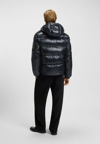 Giubbotto imbottito blu navy con esterno lucido, dotato di cappuccio e design a righe orizzontali, indossato sopra pantaloni neri e scarpe.