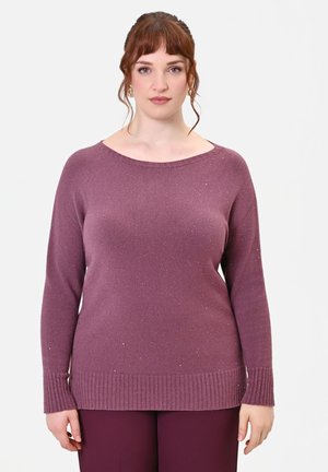 Donna con capelli rossi che indossa un maglione viola a maniche lunghe lavorato a maglia e pantaloni abbinati, in piedi davanti a uno sfondo bianco uniforme.