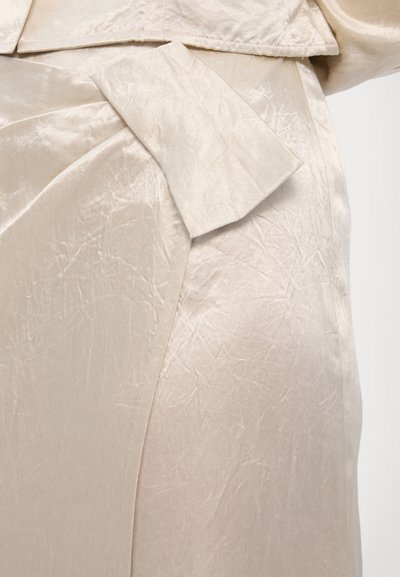Silk blouse in a light beige tone featuring a textured surface, wide collar, and asymmetric bow detailing at the waist, minimalistic design.