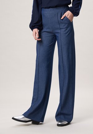 Blauwe, breed uitlopende broek met krijtstrepen, gedragen met zilveren puntige schoenen en een marineblauw blouse, met één hand in de zak, staand op een beige vloer.