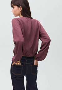 Bordeauxfarbige Bluse mit Muster, langen, puffigen Ärmeln und gerafftem Saum, kombiniert mit dunklen Jeans aus Denim mit kontrastierenden Nähten.