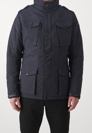 Man draagt een donkerblauwe jas met vier voorzakken, waarvan twee met ritsen op de borst en twee met knopen nabij de taille, staand.