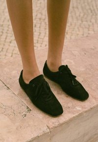 Sneaker in suede nero con punta arrotondata, lacci e piccoli fori per i lacci, appoggiati su una superficie di pietra chiara.