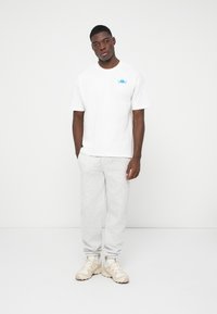 Man die tegen een witte muur staat, gekleed in een wit T-shirt met een blauw logo, lichtgrijze sweatpants en beige sneakers met witte sokken.
