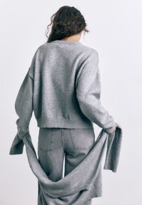 Kvinde med krøllet brunt hår iført grå sweater og jeans, der holder en anden grå sweater bag ryggen mod en ensfarvet baggrund.