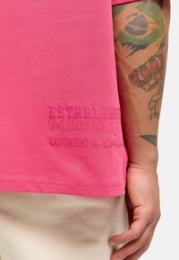Lyserød bomulds-t-shirt med præget tekst "ETABLERT Milano 1974 COPYRIGHT @ [brand]" nær kanten, glat tekstur og afslappet pasform.