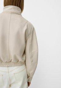 Beige cropped jacket with a high collar, elastic waistband, and subtle stitching details, made from smooth fabric.