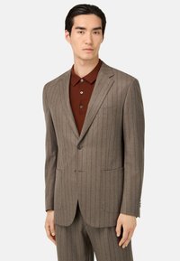 Completo marrone chiaro a righe con chiusura a due bottoni, revers a punta e tasche frontali; abbinato a una camicia marrone con colletto sotto.