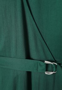 Σκούρο πράσινο φόρεμα τύπου wrap από ύφασμα με ταιριαστή ζώνη περασμένη από μια ασημένια πόρπη D-ring στη μέση.