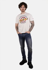 T-shirt bianca in cotone con una stampa colorata di un design circolare di un aereo e il testo "Aeronautica Militare". Abbinata a jeans blu e sneakers nere.