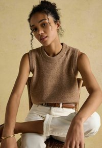 Brauner ärmelloser Strickpullover mit ribbedem Ausschnitt, kombiniert mit gerollten weißen Jeanshosen und einem braunen Ledergürtel, sitzend auf einem Holzstuhl.