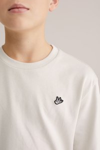 Világosszürke pamutpóló környakkal és egy kis fekete, hímzett mintával a bal mellrészen. Puha, sima tapintású anyag.