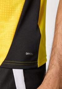 Camisa deportiva de manga corta que presenta una parte superior amarilla con un patrón texturizado y una sección inferior negra; incluye una etiqueta "DRYCELL".