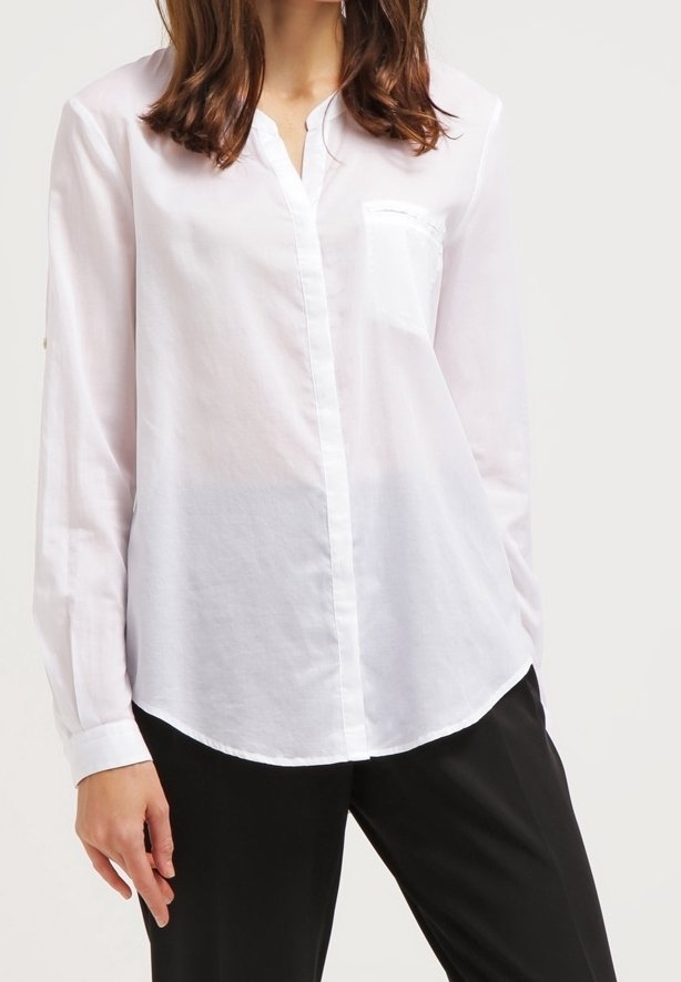 Femme portant une chemise blanche à manches longues avec une poche poitrine et un pantalon noir, debout avec une main détendue le long du corps.