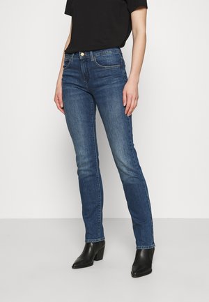 Personne portant un jean slim bleu, un haut noir à manches courtes et des bottines noires à talons, debout devant un fond uni.