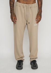 Beige sweatpants with an elastic waistband and drawstring. Side pockets and a small white logo on the thigh. Textured, soft fabric.