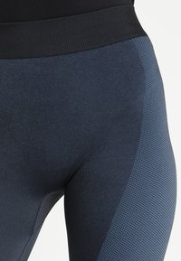 Tmavě modré legíny s texturovaným designem, s širokým černým pasem a jemnými síťovinovými akcenty po stranách. Hladký povrch látky.