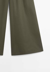 Pantalon à jambes larges en tissu vert olive avec des surpiqûres visibles à l'ourlet sur fond blanc.