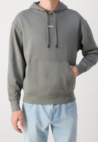 Felpa grigia con tasca a marsupio, cordoncino regolabile e logo. Tessuto morbido, polsini e orlo a coste. Indossata con jeans azzurri chiari.