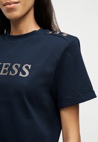 Γυναίκα που φοράει μπλε κοντομάνικη μπλούζα με τρία διακοσμητικά κουμπιά στον ώμο και μερικώς φωτεινή επιγραφή "ESS" στο μπροστινό μέρος.