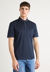 Polo azul-marinho feito de tecido suave com colarinho e uma lista de botões. Apresenta um pequeno logótipo em relevo no peito. Mangas curtas.