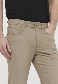 Chinos en coton beige à coupe slim. Les caractéristiques incluent une fermeture à bouton, des poches avant et des détails de couture subtils. À associer avec un haut noir.