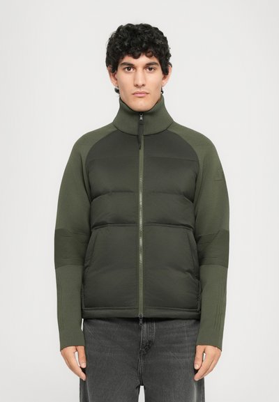 Junger Mann mit lockigem schwarzem Haar, der eine dunkelgrüne gepolsterte Jacke mit gestrickten Ärmeln und schwarze Jeans trägt, steht vor einem schlichten Hintergrund.
