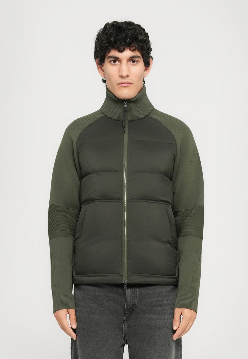 Young man with curly black hair wearing a dark green padded jacket with knitted sleeves and black jeans, standing against a plain background.