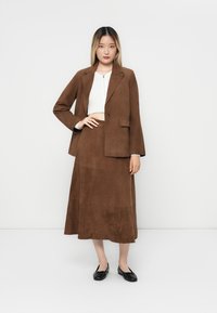 Blazer en daim marron et jupe assortie, forme en A, texture lisse, détail de bouton sur le blazer, associé à un haut blanc et des ballerines noires.