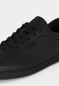 Baskets basses en cuir noir avec lacets noirs, surface texturée et logo "GUESS" embossé sur le côté et la languette.