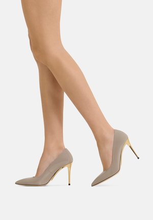 Gambe che indossano décolleté beige con punta appuntita e tacchi a spillo dorati, in piedi su sfondo bianco.