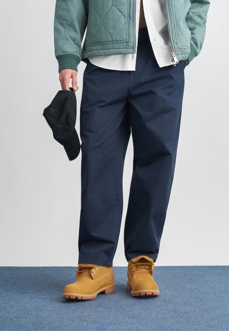 Granatowe, luźne spodnie w połączeniu z zieloną pikowaną kurtką, białą koszulą i beżowymi butami roboczymi. W jednej ręce trzymana jest czarna czapka.