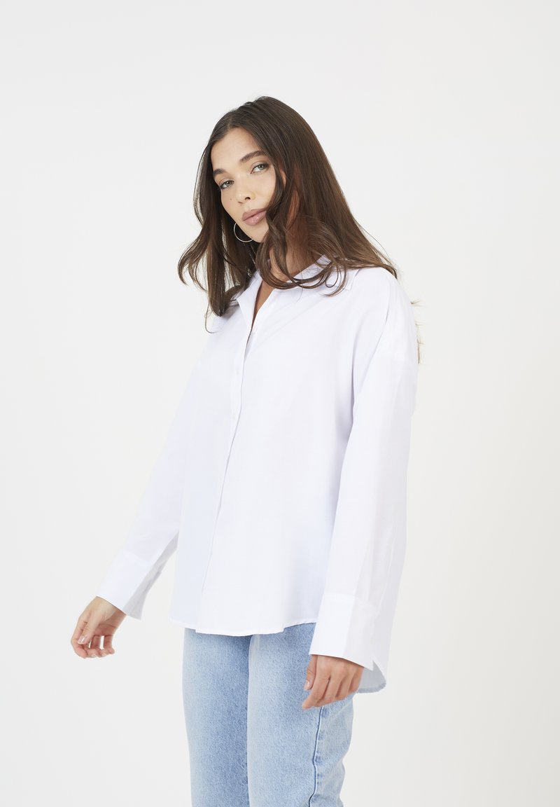 Giovane donna con lunghi capelli castani, che indossa una camicia bianca larga con bottoni e pantaloni in jeans azzurri chiari, in piedi davanti a uno sfondo bianco uniforme.