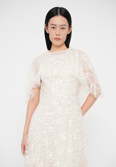 Ένα ανοιχτό μπεζ κεντητό φόρεμα με λουλουδάτα σχέδια, διάφανα φαρδιά μανίκια και εφαρμοστή μέση με αέρινη φούστα.