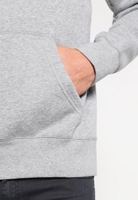 Sudadera gris hecha de material suave, con un bolsillo frontal, puños acanalados y un corte relajado. Se pueden ver detalles de costura cerca del dobladillo.