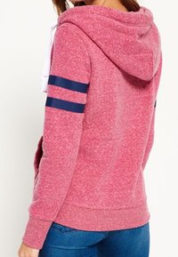 Donna che indossa una felpa con cappuccio rosa con due strisce blu navy sulle maniche e jeans blu, vista da dietro.
