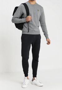 Sudadera gris con un logo amarillo, combinada con pantalones de chándal negros y zapatillas blancas. Presenta un diseño simple y casual y una mochila ajustable.