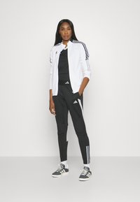 Zwart-witte Adidas trainingsoutfit met een ritsjack met drie strepen op de mouwen, bijpassende taperende broek en grijze sneakers.