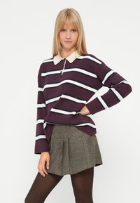 Jauna sieviete ar taisniem blondiem matiem, tērpta purpura un baltā svītrainā garās piedurkņu polo krekla, svītrainā svārku un melnām zeķbiksēm, stāv ar roku uz gurniem.
