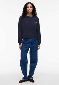 Jūrų laivyno džemperis su baltais "Niujorkas 1994" siuvinėjimais, derinamas su šviesiai mėlynomis, laisvo kirpimo džinsais ir rudais basakojais.