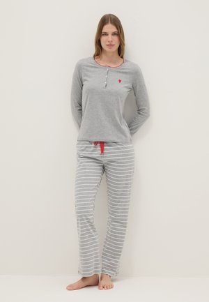 Frau trägt ein graues Langarmshirt mit rotem Herz-Detail und grau-weiß gestreifte Pyjamahose mit rotem Kordelzug, steht barfuß.