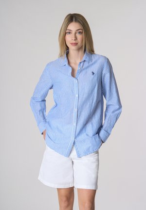 Camicia a righe blu e bianche con colletto, maniche lunghe e un piccolo simbolo ricamato sul petto. Abbinata a pantaloncini bianchi.