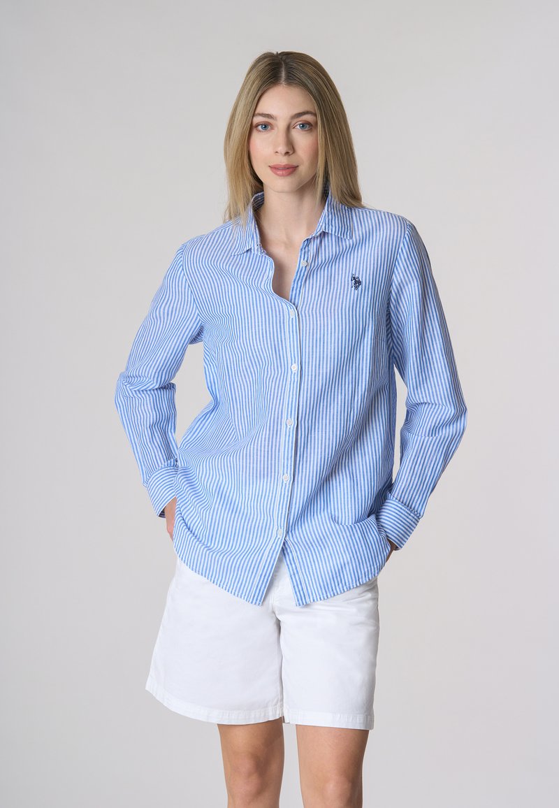 Camicia a righe blu e bianche con colletto, maniche lunghe e un piccolo simbolo ricamato sul petto. Abbinata a pantaloncini bianchi.