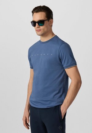 Mann trägt blaues Hackett-T-Shirt und dunkle Hose mit einer Hand in der Tasche, trägt schwarze Sonnenbrille, vor einfachem hellem Hintergrund.
