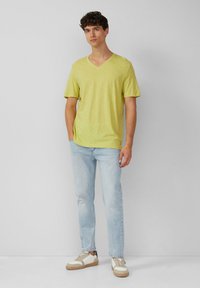 V-hals, t-shirt met korte mouwen in helder geel, gecombineerd met lichtblauwe jeans en witte sneakers met tan accenten. Gladde textuur, losse pasvorm.