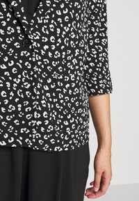 Blazer à imprimé léopard noir et blanc avec un tissu texturé, fermeture à un bouton, et manches trois-quarts. Dispose d'une poche plaquée.