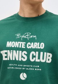 Πράσινο μπλουζάκι με στρογγυλή λαιμόκοψη και λευκό κείμενο που γράφει "Monte Carlo Tennis Club" και "Health and Sports Club established by Björn Borg" με λογότυπο μπάλλας τένις.
