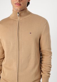 Pull beige zippé avec un col haut, texture côtelée et petit logo avec des accents rouge, blanc et bleu sur la poitrine.