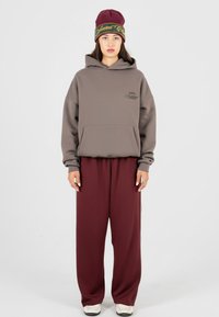 Hoodie gris surdimensionné avec poche frontale et logo, associé à un pantalon bordeaux à jambes larges. Le mannequin porte un bonnet marron avec des accents verts.