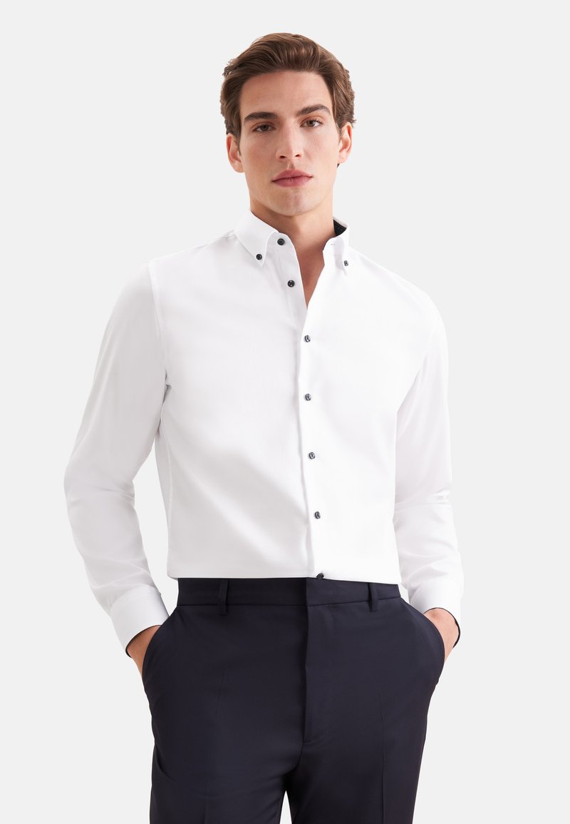 Jeune homme portant une chemise blanche boutonnée et un pantalon sombre, debout avec les mains dans les poches devant un fond uni.